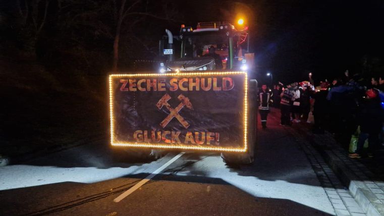 Ein beleuchteter Traktor mit einem Schild, das "ZECHE SCHULD GLÜCK AUF!" zeigt. Im Hintergrund sind Menschen auf der Straße bei Nacht zu sehen.