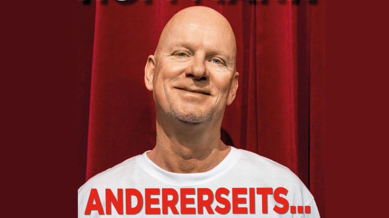 A smiling man stands in front of a red curtain. He is wearing a white T-shirt with a striking print.