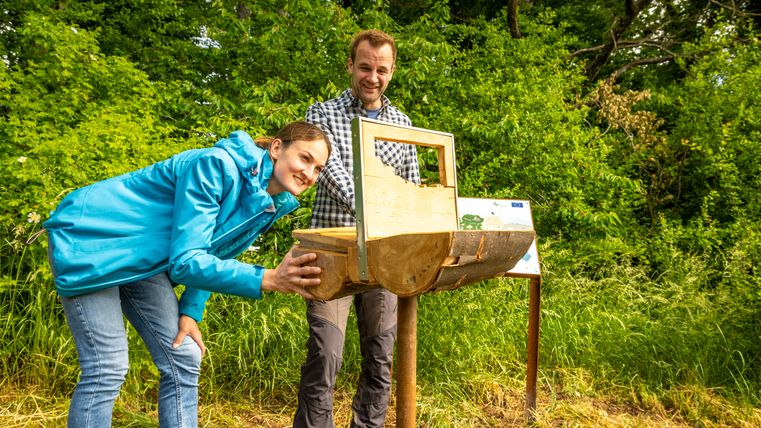 Twee mensen kijken naar een houten object in een groene omgeving.