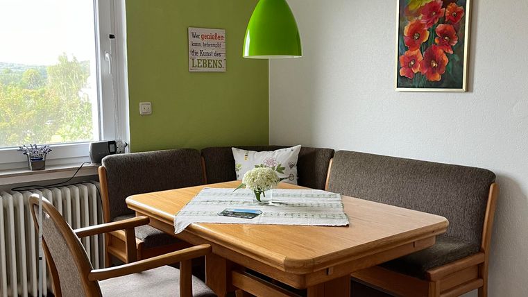 Ein gemütlicher Essbereich mit einem Holzstisch und beigen Polstermöbeln. An der Wand hängt ein Bild mit roten Blumen und eine grüne Lampe hängt von der Decke.