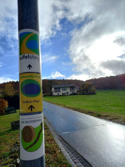 Ein Wegweiser an einem Pfosten zeigt verschiedene Wanderwege in der Eifel an. Im Hintergrund ist ein Haus und eine grüne Wiese unter einem bewölkten Himmel zu sehen.
