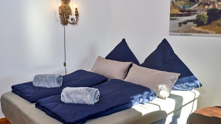 Ein gemütliches Schlafzimmer mit einem großen Bett und blauen Bettdecken. Im Hintergrund ist ein Wandbild und eine dekorative Lampe zu sehen.