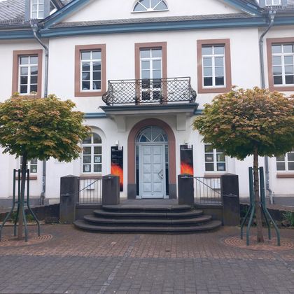 Een historisch gebouw met een blauwe voordeur en symmetrische ramen. Voor het gebouw staan twee gelijkmatig gesnoeide bomen.