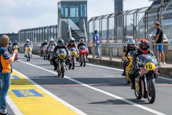 Een groep motorfietsen rijdt op het circuit. Toeschouwers kijken vanaf de zijlijn naar de gebeurtenissen.