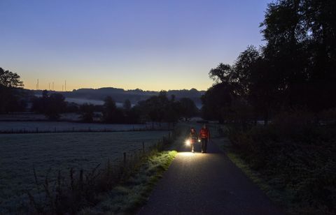 Zwei Personen gehen auf einem Weg in der Dämmerung, während sie eine Taschenlampe tragen. Die Umgebung ist frostig und die Landschaft wirkt ruhig und friedlich.