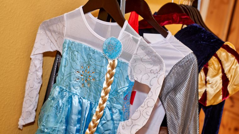 Kostüme für Kinder hängen an einem Kleiderbügel. Darunter befindet sich ein zauberhaftes blaues Kleid mit einer langen Zopfperücke.