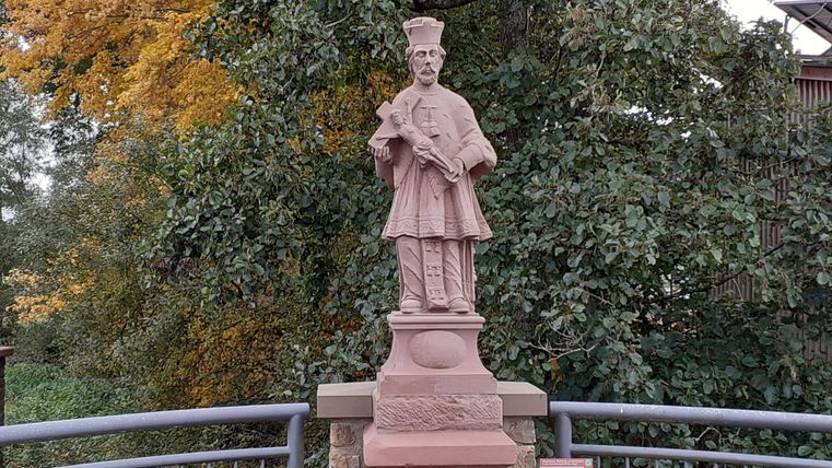 Standbeeld van St. Johannes van Nepomuk op een brug, omringd door herfstbomen.