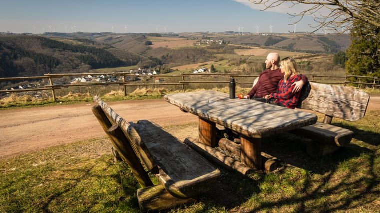 Un couple est assis sur un banc à une table en bois avec vue sur un paysage vallonné et des éoliennes au loin.