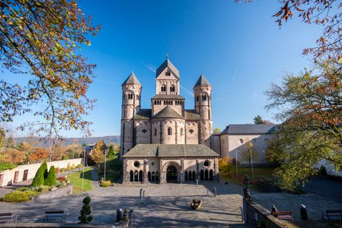 Une église imposante avec des tours et un ciel bleu clair. Au premier plan, on voit une place entourée d'arbres.