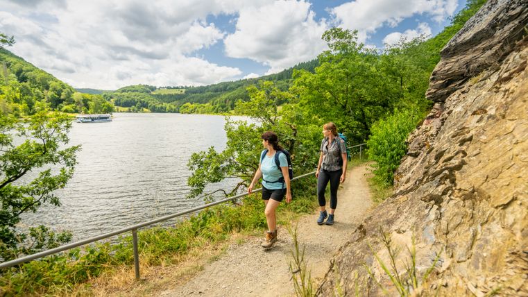 Zwei Wandererinnen auf einem schmalen Pfad am Rursee, der sich entlang eines felsigen Hangs schlängelt. Umgeben von üppigem Grün und bewaldeten Hügeln genießen sie die Aussicht auf das ruhige Wasser unter einem teils bewölkten Himmel