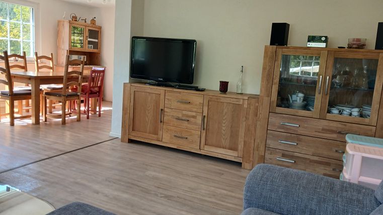 Ein gemütliches Wohnzimmer mit einem Fernseher und Holz Möbeln. Im Hintergrund ist ein Essbereich mit einem großen Tisch und Stühlen zu sehen.