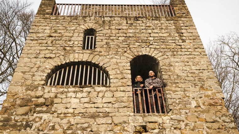 Twee mensen staan op een balkon van een oud stenen kasteel met tralievensters.