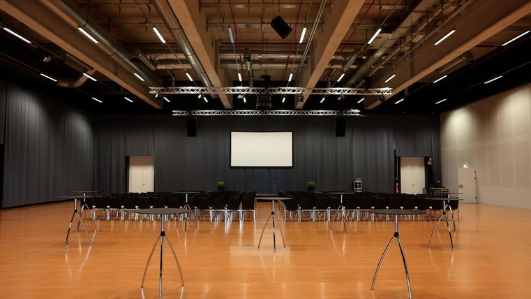 A large event space with wooden floors and modern lights. In the center are rows of chairs and a large screen for presentations.