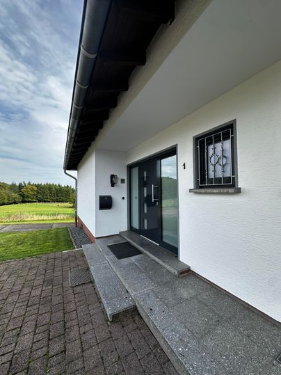 Une entrée moderne avec une porte grise et de grandes fenêtres. Le chemin est pavé et mène à un espace vert bien entretenu.