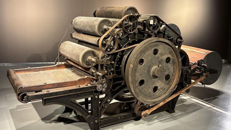 Een oude drukmachine met meerdere rollen van metaal en hout. Ze staat in een moderne tentoonstellingsruimte.
