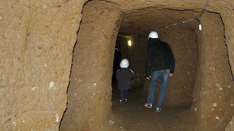 Eine schmale, erdige Höhle mit zwei Personen, die einen Helm tragen. Das Licht am Ende der Höhle schafft eine spannende Atmosphäre.