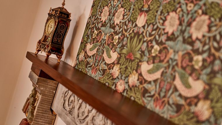 Ein eleganter Kamin mit einem dekorativen Holzregal. Im Hintergrund ist ein kunstvolles Ornament und eine antike Uhr sichtbar.