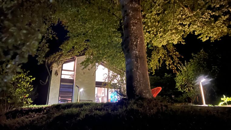 Ein modernes Haus in der Nacht, umgeben von Bäumen. Weiches Licht beleuchtet den Außenbereich und schafft eine gemütliche Atmosphäre.