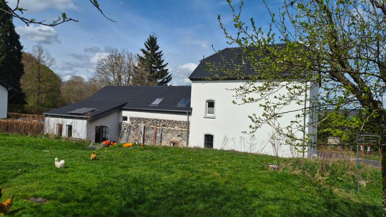 Een groot, modern huis met een schilddak en een aangelegde grasveld aan de voorkant. Op de achtergrond zijn bomen en een heldere lucht zichtbaar.