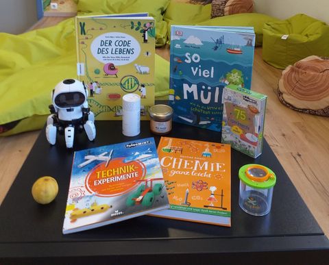 A table with various books, including titles on technology and chemistry. Additionally, there is a small robot, a glass, and an apple visible.