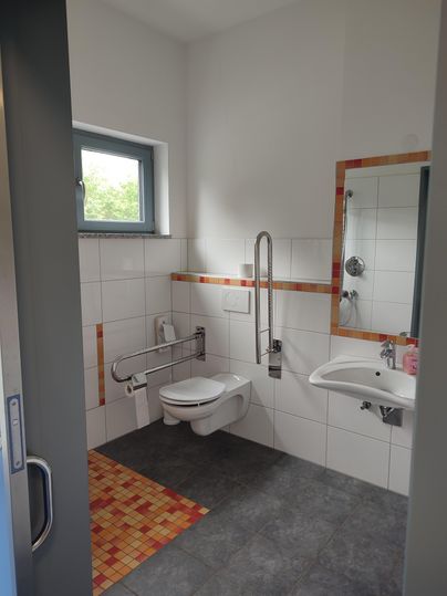 Une salle de bain moderne avec des carreaux blancs et un accès pour les personnes handicapées. Il y a un lavabo, des toilettes et une douche.
