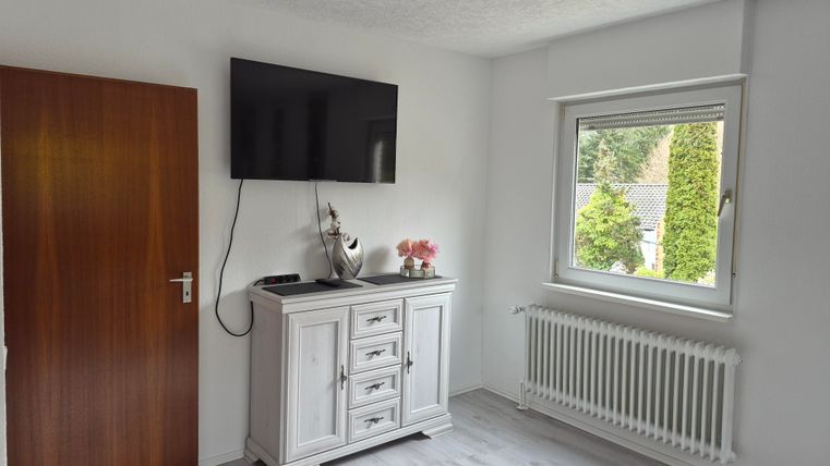 Een lichte kamer met een televisie aan de muur en een wit dressoir. Het raam biedt uitzicht op de groene omgeving.