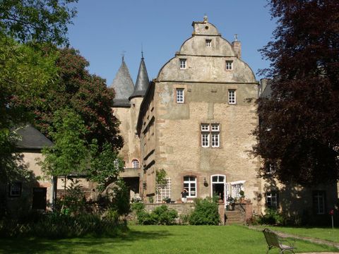 Historical building with towers, surrounded by trees and a well-kept garden. A terrace is visible, the weather is sunny.
