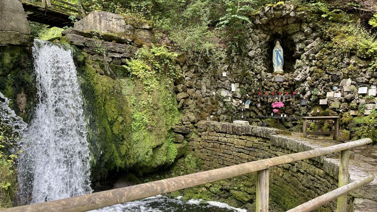 Een schilderachtige bron met waterval, omgeven door groene planten. Aan de rotswand bevindt zich een standbeeld van Maria, omgeven door bloemen.