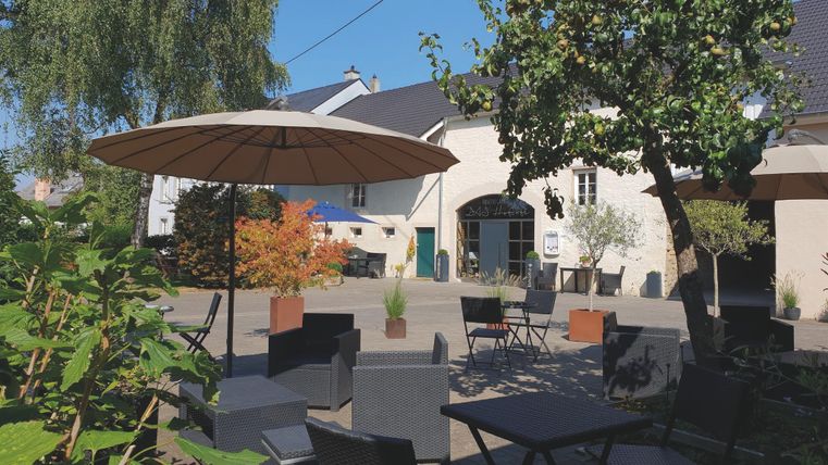 Ein gemütlicher Hof mit Tischen, Stühlen und Sonnenschirmen, umgeben von Bäumen und Pflanzen, vor einem Gebäude mit der Aufschrift 'Hofcafe Messerich'.