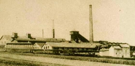 Une ancienne installation industrielle avec de grandes cheminées et plusieurs bâtiments. Les environs sont ruraux et vastes.