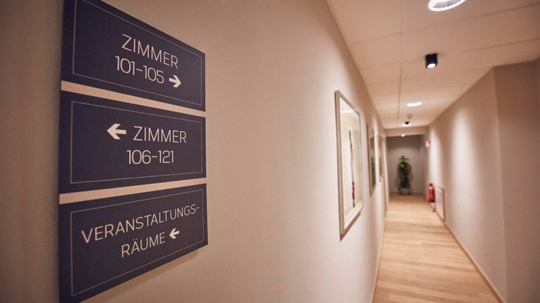 Een lichte gang met bewegwijzering naar verschillende kamers en vergaderruimtes. De vloer is van hout en de muren zijn neutraal geschilderd.