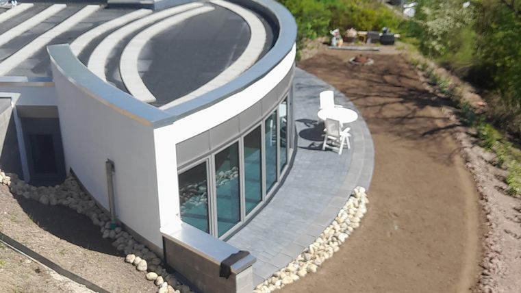 Een modern huis met een gebogen gevel en grote ramen. Omgeven door een groen landschap en een aangelegde tuin.