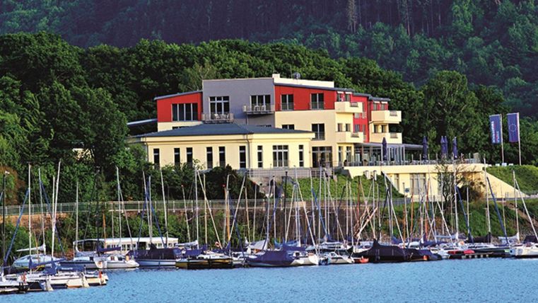 A modern building by the water with many sailboats in the foreground. The surroundings are green and offer a beautiful view of nature.