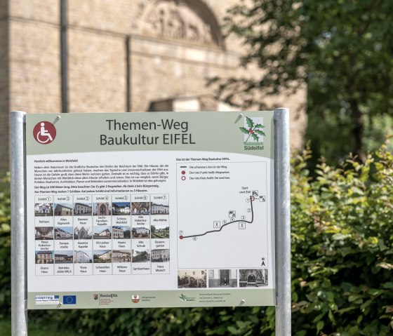 Schild des Themenwegs Baukultur Eifel in Wolsfeld mit Karte und Informationen zu Sehenswürdigkeiten. Im Hintergrund ist ein Gebäude zu sehen., © Naturpark Südeifel, Thomas Urbany