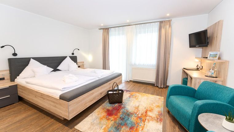 Een moderne gastenkamer met een groot bed en een comfortabele stoel. De kamer is licht en vriendelijk, met een kleurrijk tapijt en een tv.
