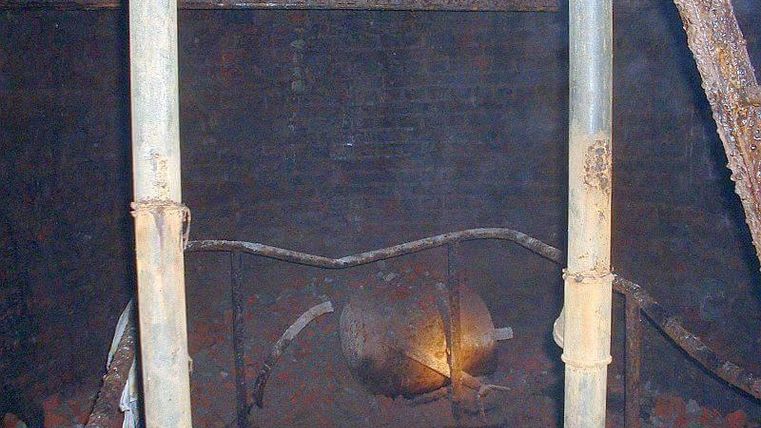 A dark room with rusty pipes and a faint light. In the background, a large, round structure is visible.