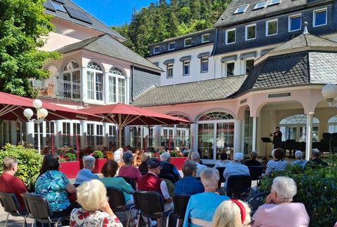 Eine schöne, sonnige Veranstaltung im Freien mit einem Publikum, das einem Musiker zuhört. Im Hintergrund sind elegante Gebäude und grüne Bäume zu sehen.
