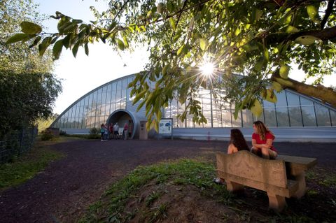 Une structure moderne en verre dans la nature. Deux femmes sont assises sur un banc et profitent du soleil.