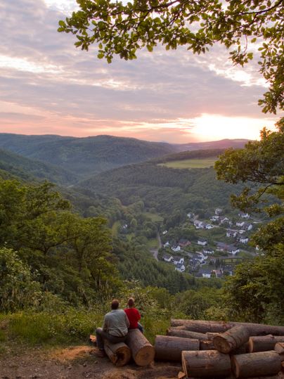 Zwei Personen sitzen auf Baumstämmen und blicken auf ein Tal mit einem Dorf, umgeben von bewaldeten Hügeln bei Sonnenuntergang.
