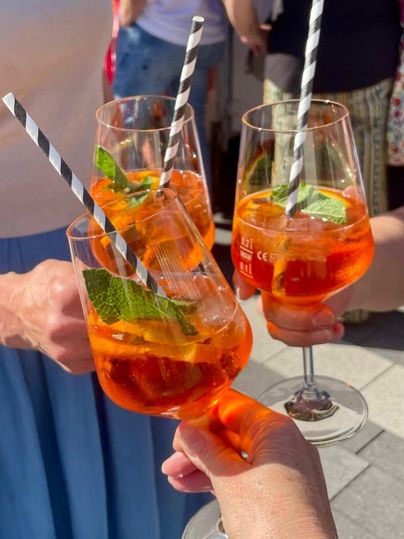 Drei Gläser mit erfrischenden Cocktails, dekoriert mit einem Minzblatt und gestreiften Strohhalmen. Im Hintergrund sind Menschen in der Sonne zu sehen.