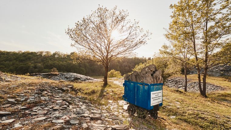 Eine mit Schiefergestein gefüllte blaue Lore steht im Sonnenlicht auf einer Halde, umgeben von lichten Bäumen und Natur; im Hintergrund dichte Wälder und ein einzelner Baum, durch dessen Äste die Sonne scheint.