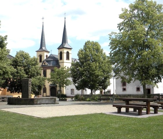 Une place de village idyllique à Idenheim avec une église en arrière-plan, entourée d'arbres. Au premier plan, on peut voir des bancs et une fontaine., © Ingrid Penning