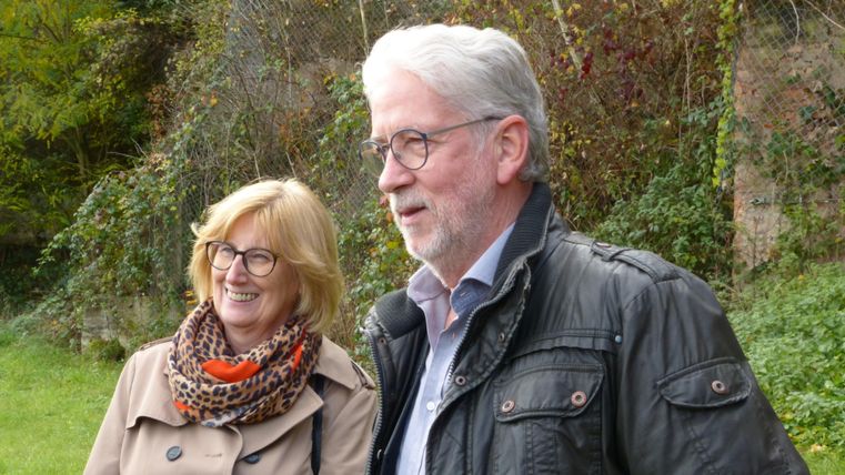 Ein fröhliches Paar steht im Freien. Im Hintergrund sind Pflanzen und Bäume zu sehen.