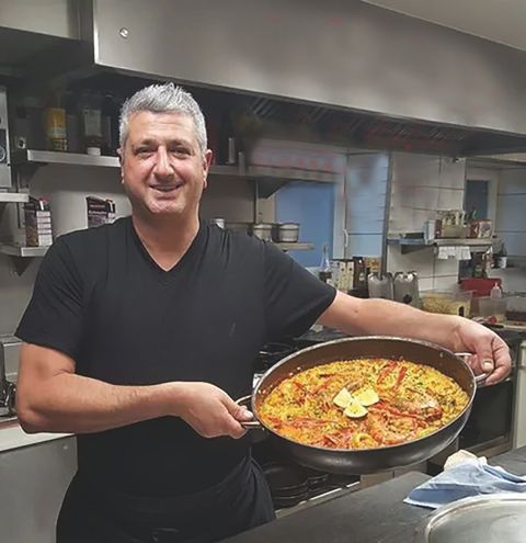 Man smiles and presents a pan with a rice dish in it; is located in a kitchen