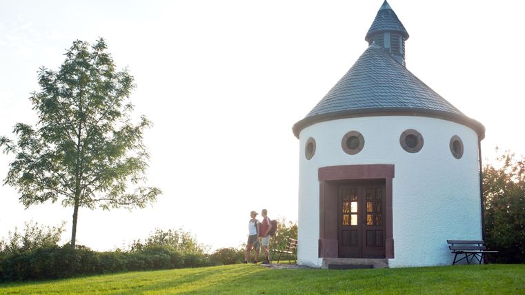 A small, round chapel stands in a green meadow. Two people are standing in front of it and enjoying the view.