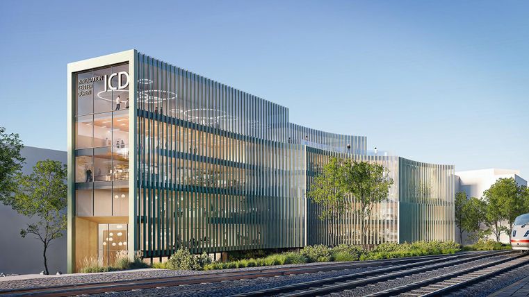 Ein modernes Gebäude mit Glasfassade und begrünten Flächen. Im Vordergrund sind Schienen für Züge zu sehen.