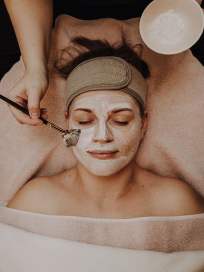 Een vrouw krijgt een gezicht behandeling met een masker. Ze ligt ontspannen op een handdoek terwijl een behandelaar het masker aanbrengt.