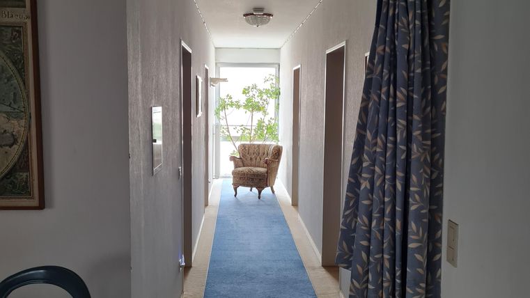 Ein schmaler Flur mit blauen Teppichboden und einem dekorativen Vorhang. Am Ende des Flurs steht ein gemütlicher Sessel.