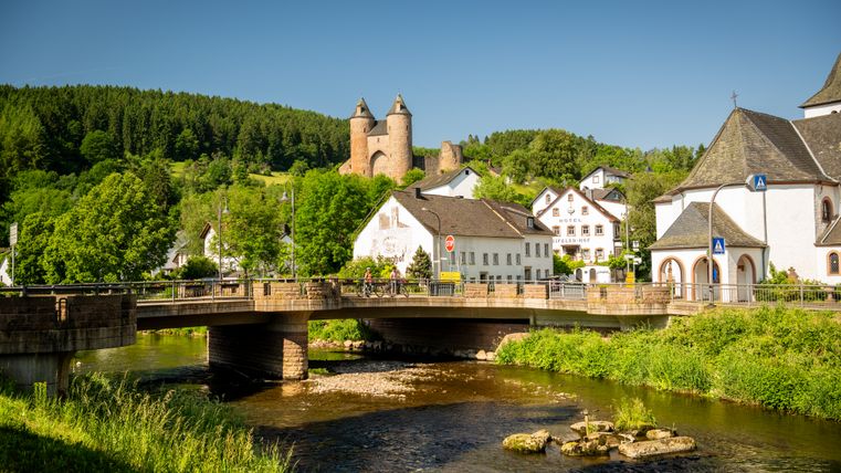 Une rivière avec un pont au-dessus. Derrière, quelques habitations et un majestueux château au-dessus.
