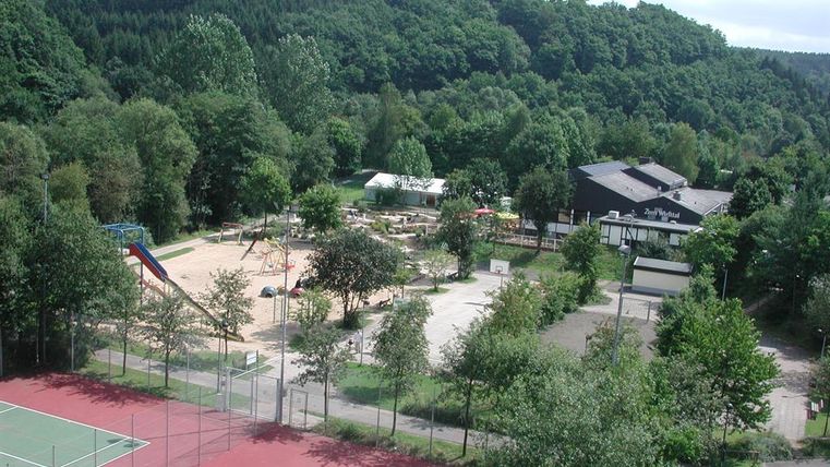 Amusement park with tennis court, playground, and restaurant in a wooded environment. Dense forests in the background, sports and play facilities in the foreground.